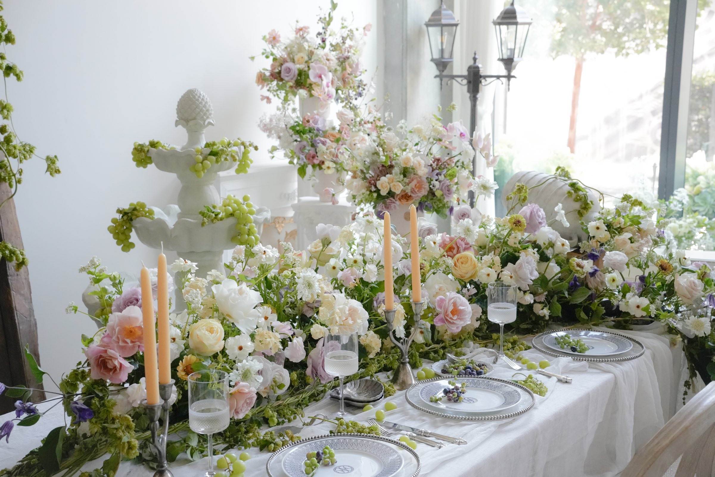 A long table styled with garden roses, ranunculus, taper candles and silver-rimmed place settings in soft daylight