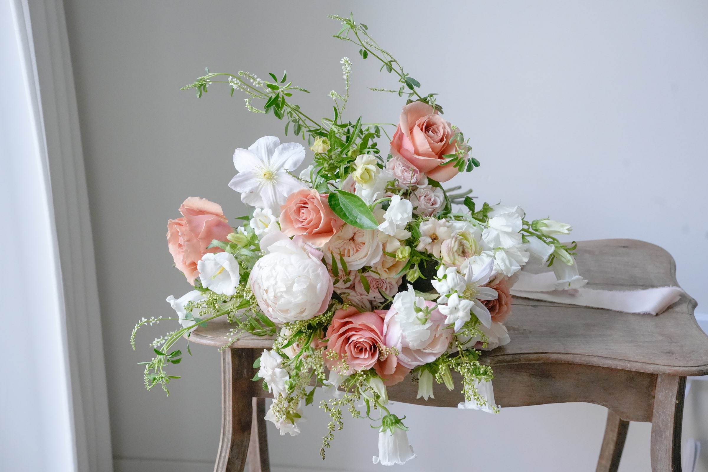 A peach and ivory bridal bouquet with garden roses, a white peony and clematis, resting on a weathered wooden stool