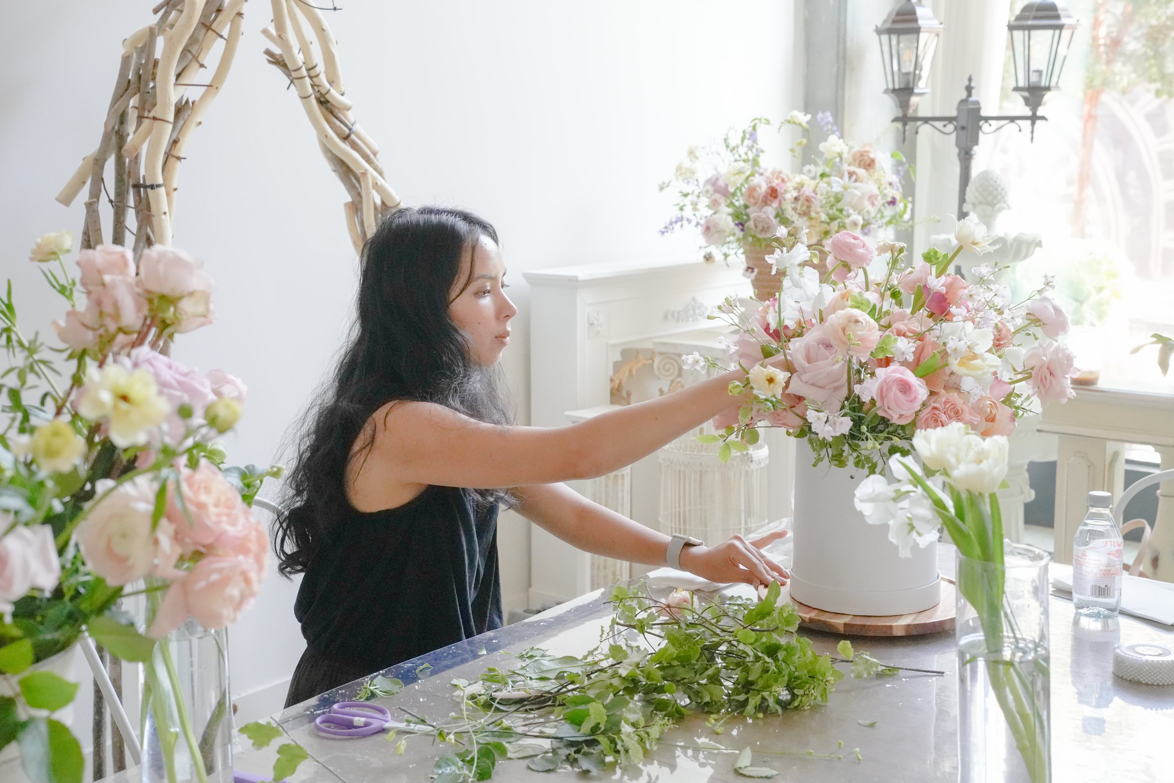 A florist arranging pink garden roses and ranunculus into a tall centerpiece at a sunlit studio table