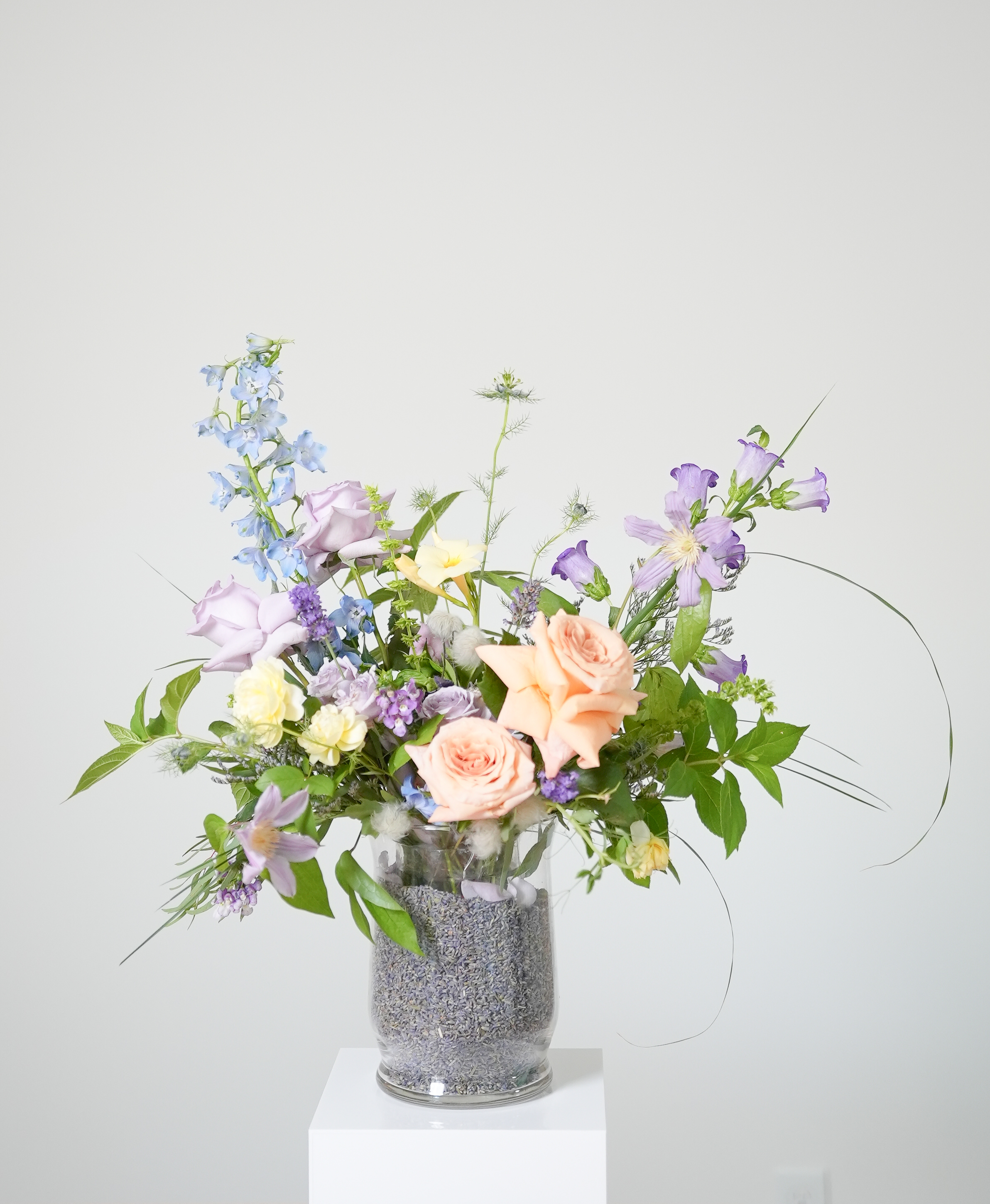 Pastel Meadow Vase — pastel roses, pale blue delphinium, purple clematis, fresh greenery, and lavender-filled glass vase